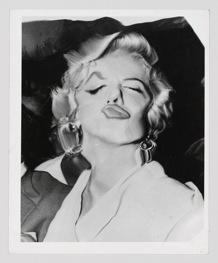 Blonde woman with closed eyes puckering her lips, wearing hoop earrings and a white blouse.