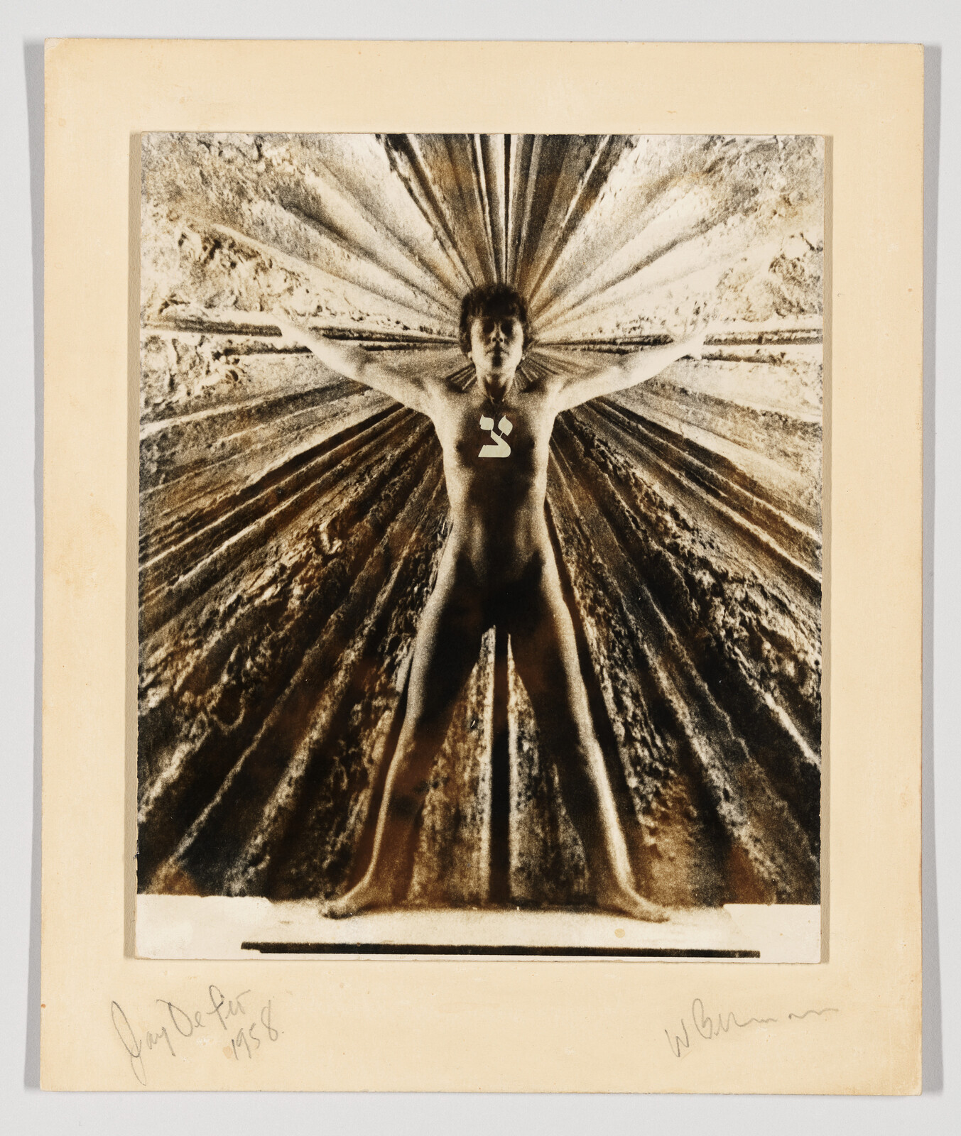 Person standing with arms and legs spread against a textured background, wearing a necklace with a symbol.