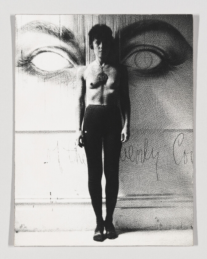 A shirtless young man stands rigidly against a wall with large painted eyes above him.