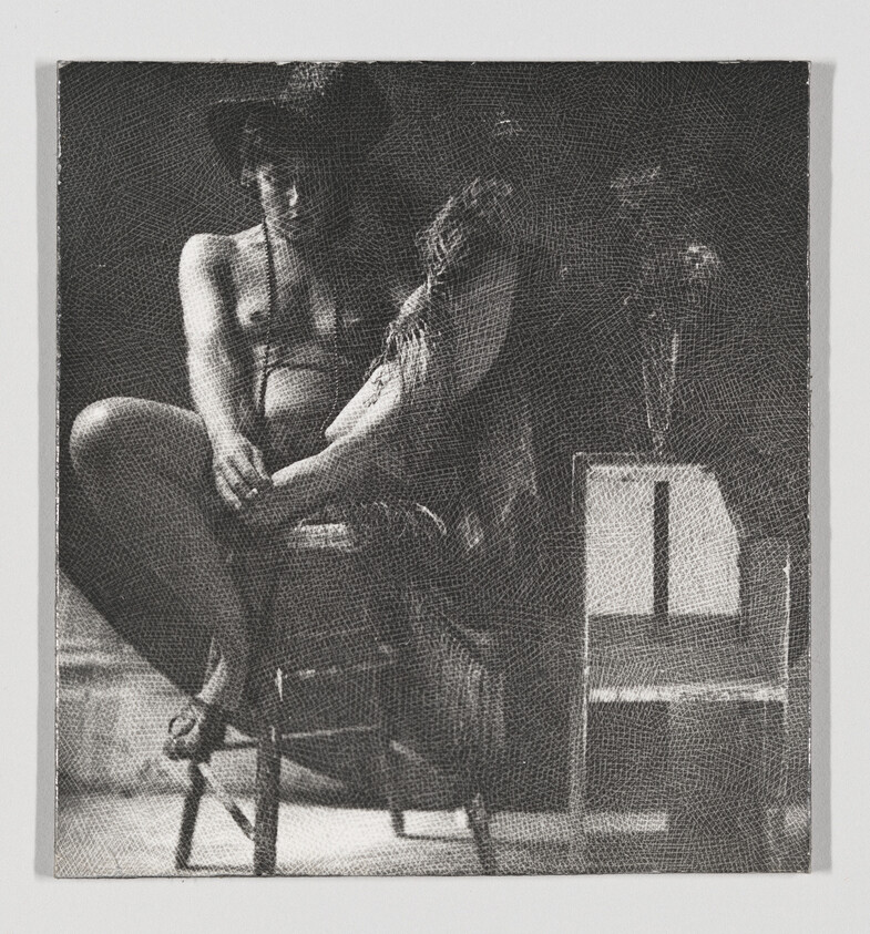 A seated woman wearing a hat and shawl sits with crossed legs on a wooden chair.