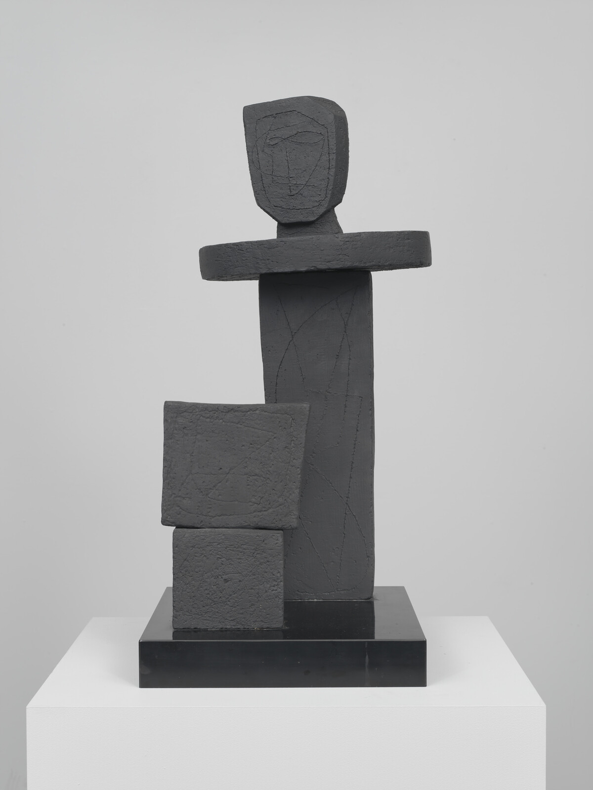 Abstract dark sculpture of stacked geometric blocks suggesting a standing figure on a white pedestal.