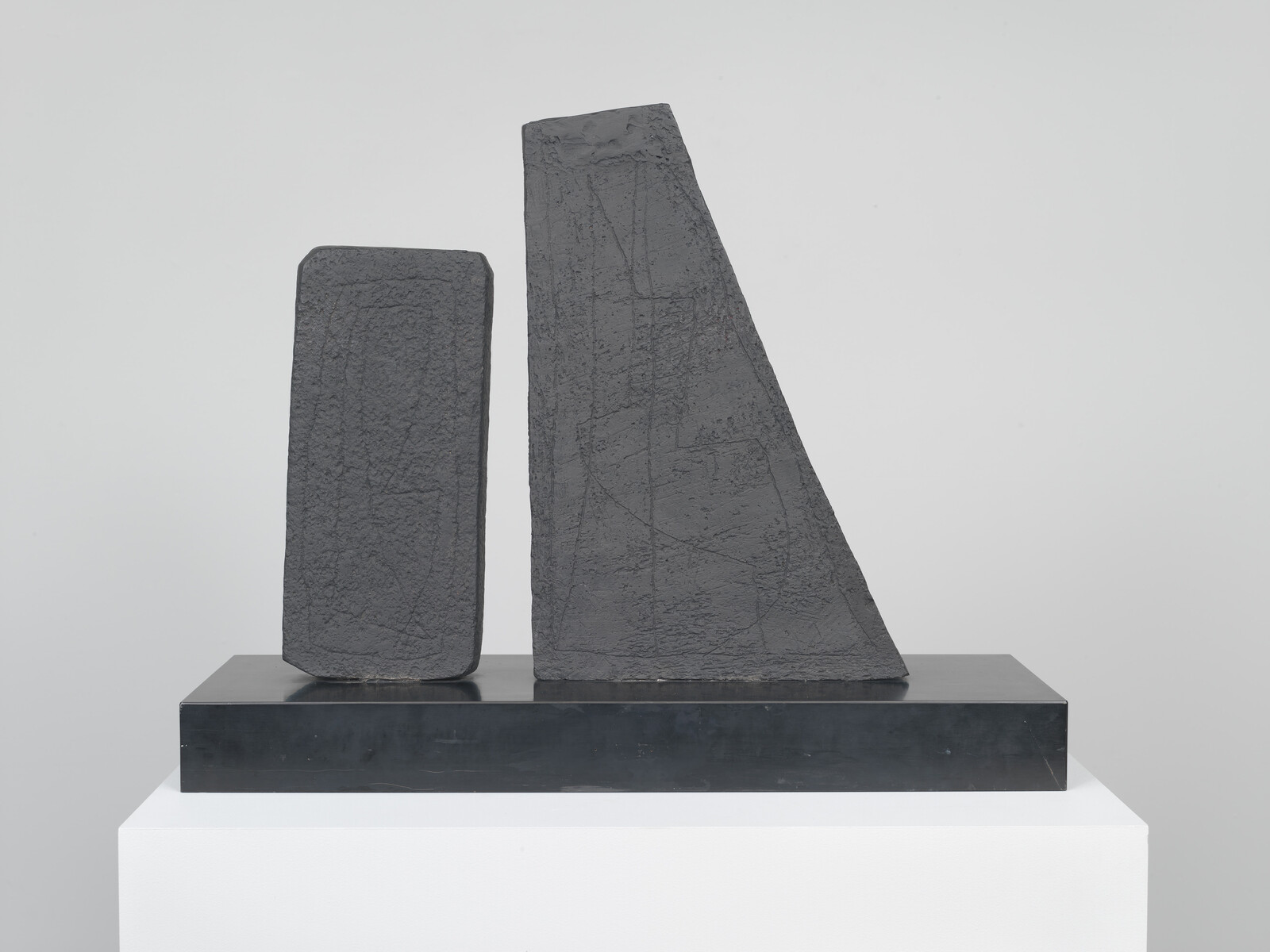 Two dark abstract stone blocks stand side by side on a black base atop a white plinth.