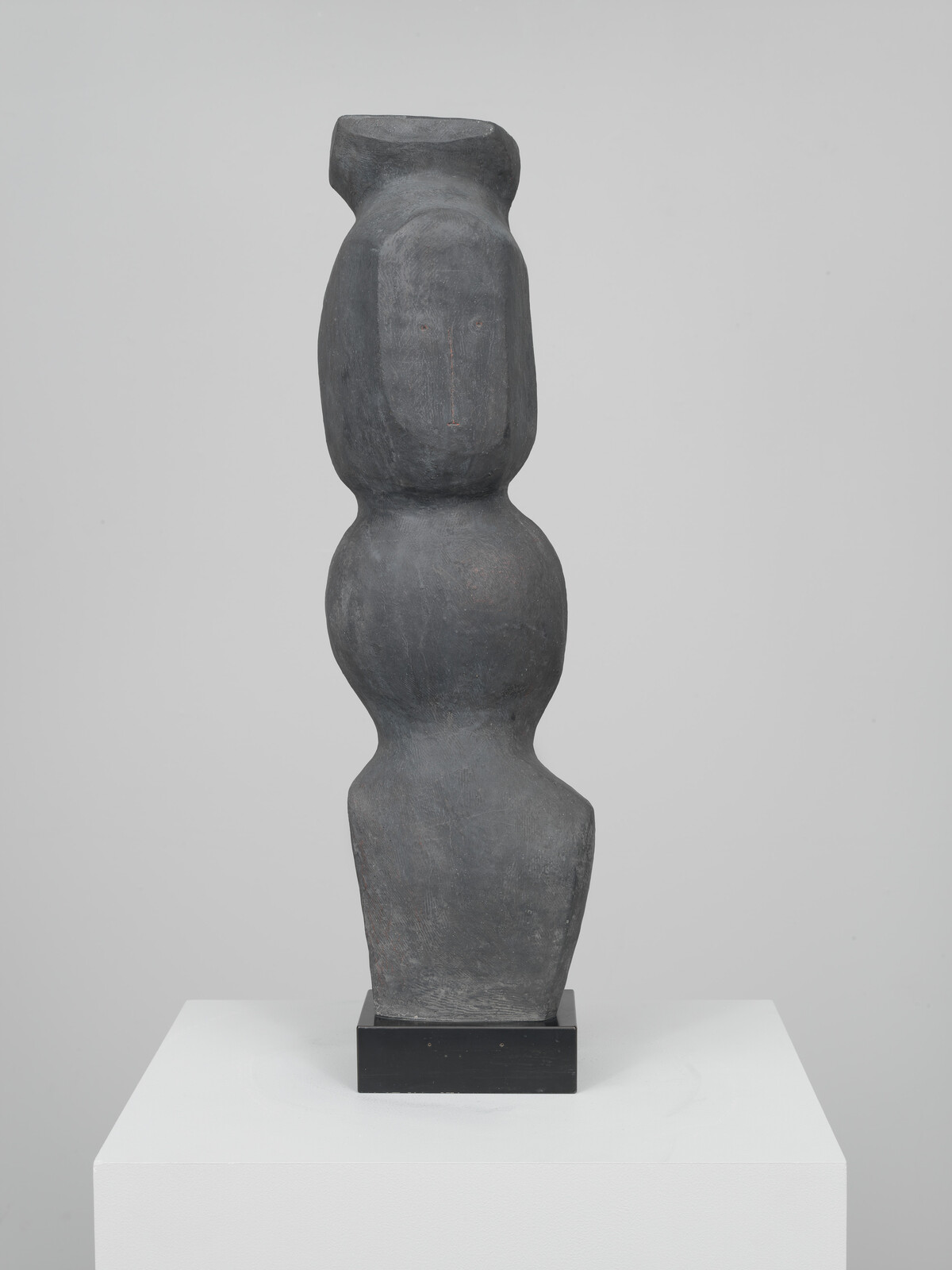 Tall dark abstract sculpture resembling a simplified standing human figure on a white pedestal.