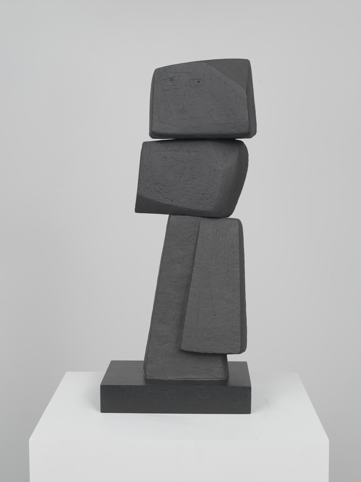 A dark geometric sculpture of stacked blocks resembling a standing figure with a faint carved face.