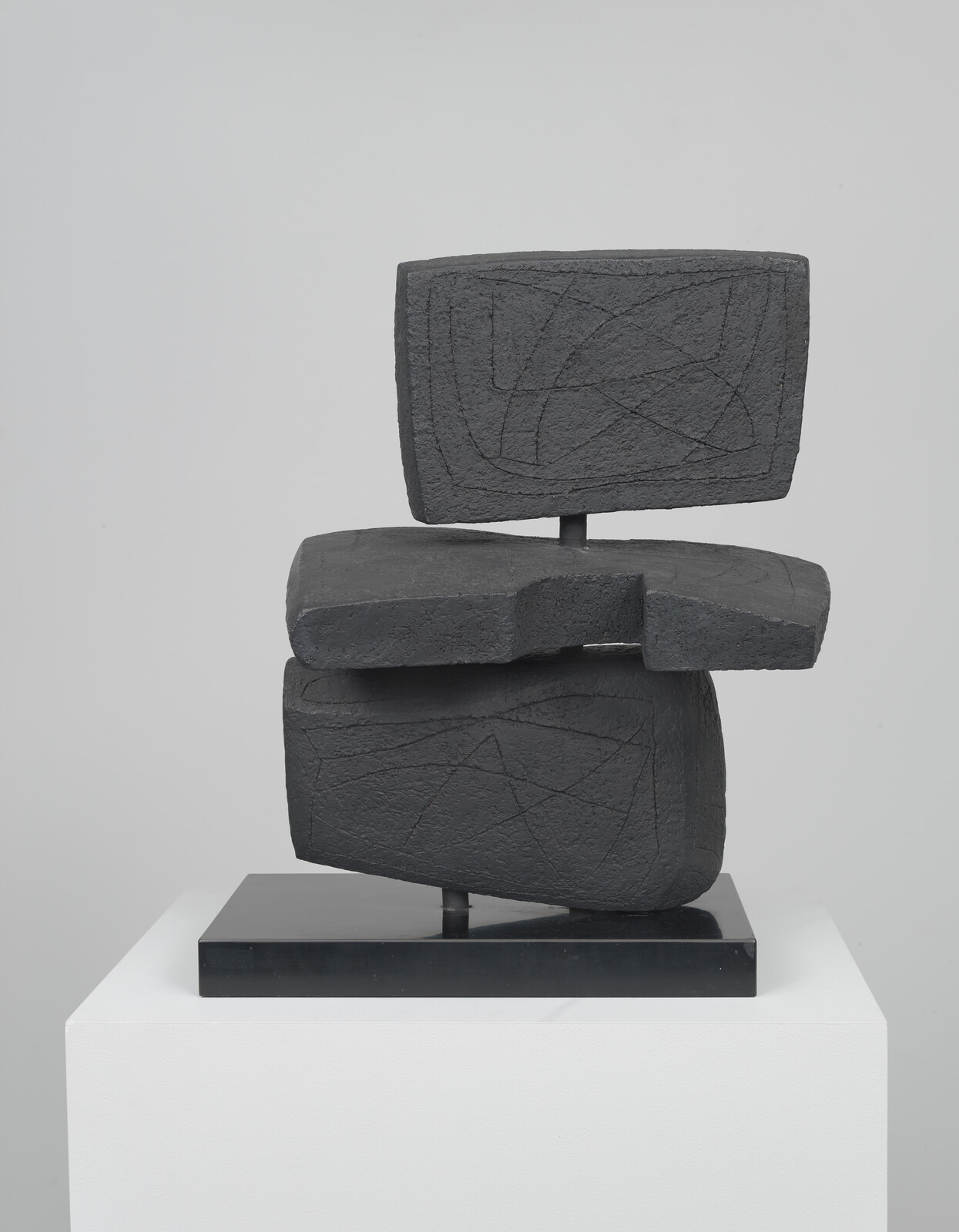 Abstract dark stone-like sculpture of three stacked blocks with carved lines on a white pedestal.