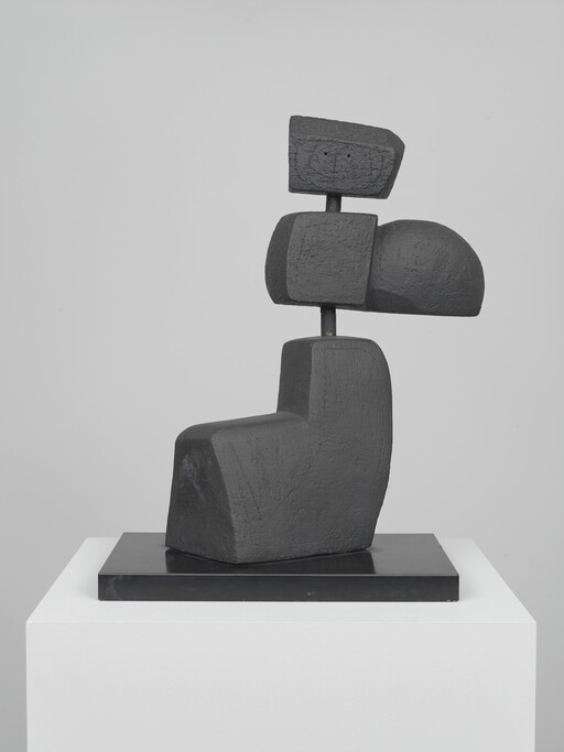 Abstract dark sculpture of stacked geometric blocks balanced on a black base atop a white pedestal.