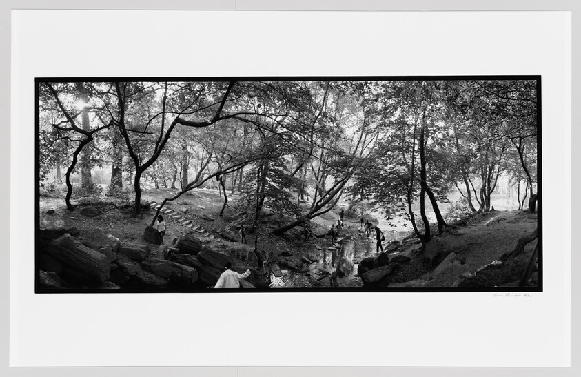 Several people walk and sit on rocks beside a shallow stream in a tree-filled park.