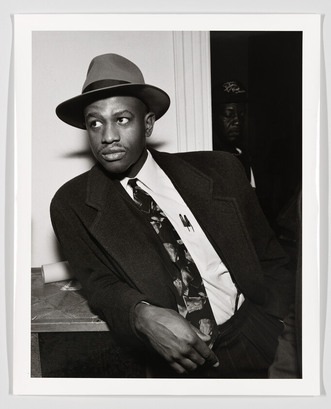 Well-dressed man in a hat and suit leans against a table, looking to the left.