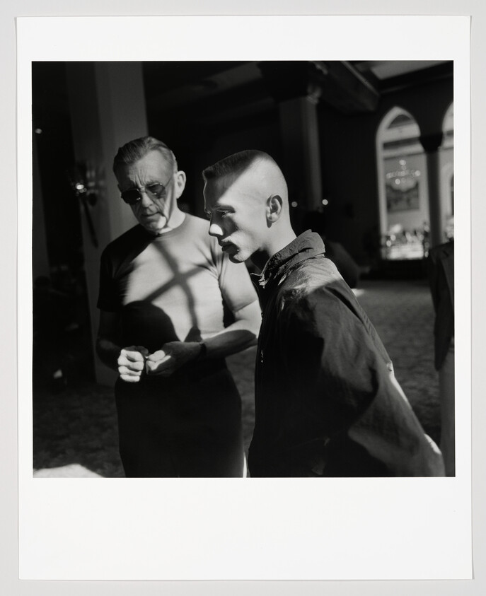 An older man and a younger man with a high-fade haircut stand and talk in strong light and shadow.