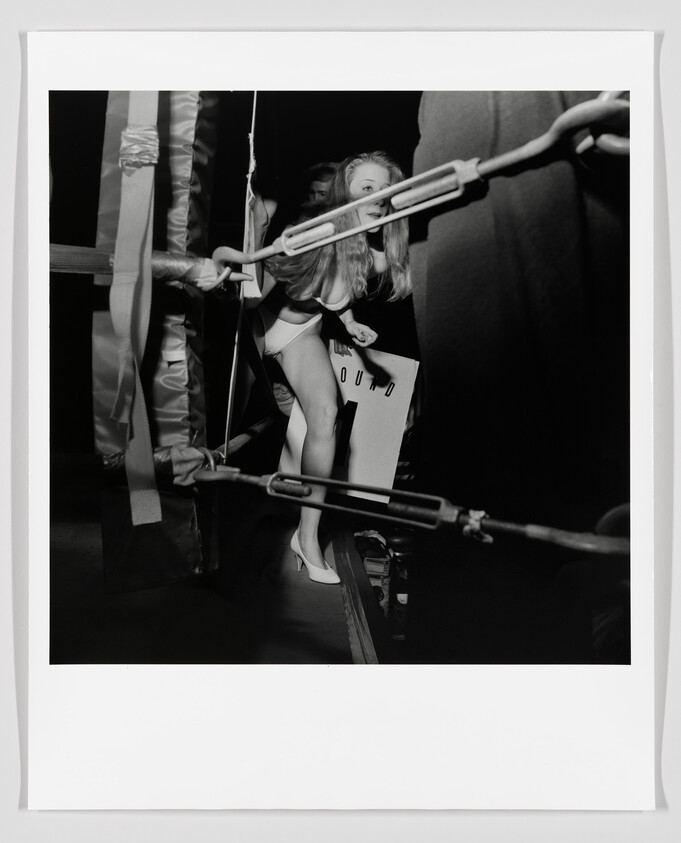 Young woman in a bikini steps through boxing ring ropes while holding a sign.