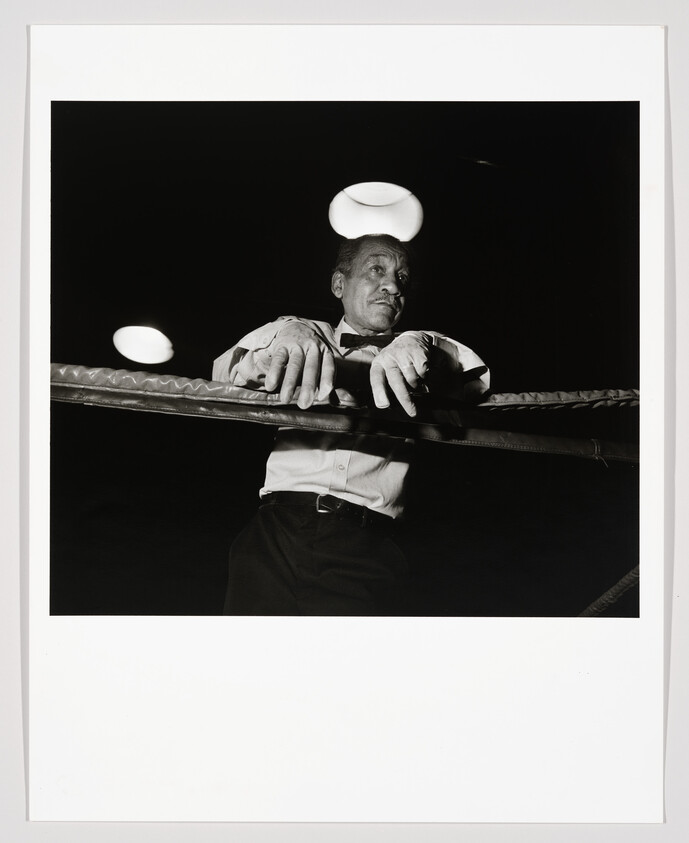 A boxing referee leans on the ring ropes under overhead lights, watching the match intently.
