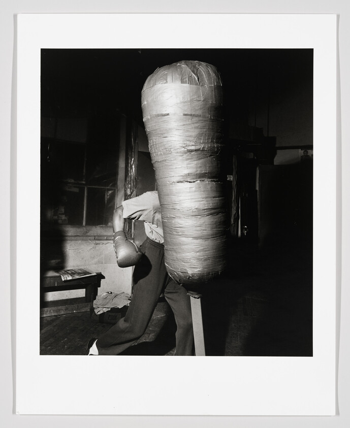 Boxer in gloves practices punches on a tall wrapped heavy bag in a dim gym.