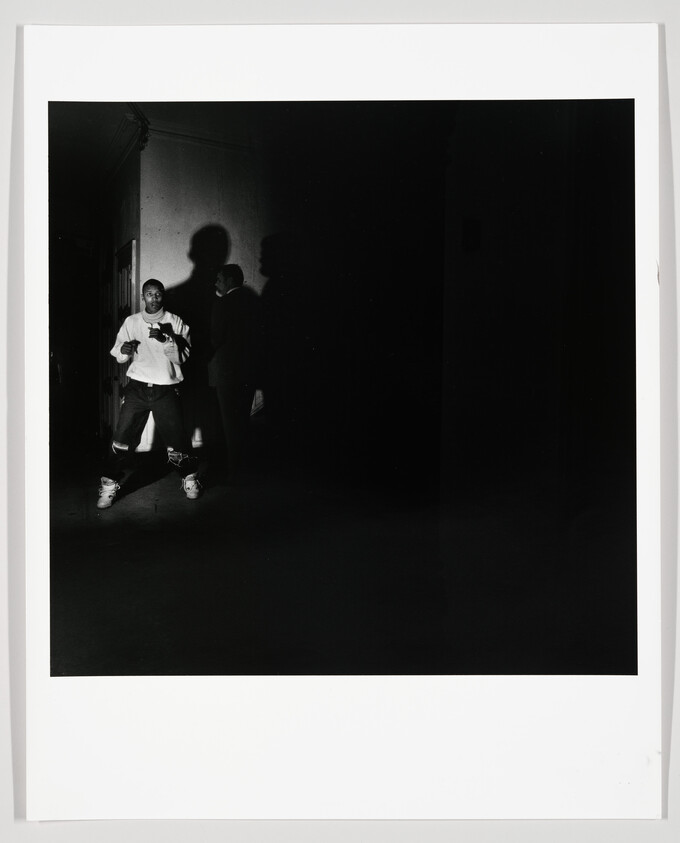 A young man stands in a dark hallway casting a large shadow while another person stands behind him.