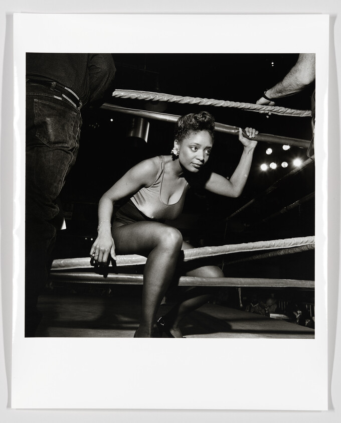 Woman in a leotard climbs through boxing ring ropes, gripping the top rope with determination.