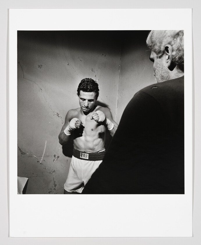 Boxer with wrapped hands stands in a corner with fists raised while trainer watches.