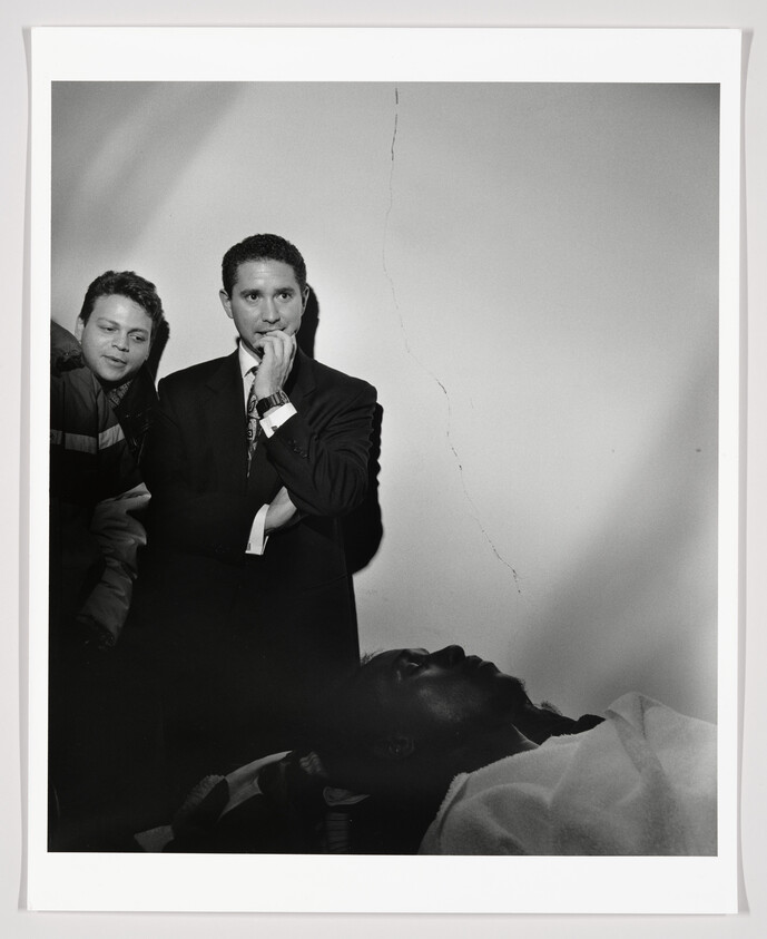 A man in a suit stands with hand to his mouth while another watches over a lying, covered person.