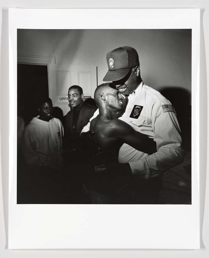 Security officer embraces a shirtless man warmly while two onlookers watch nearby.