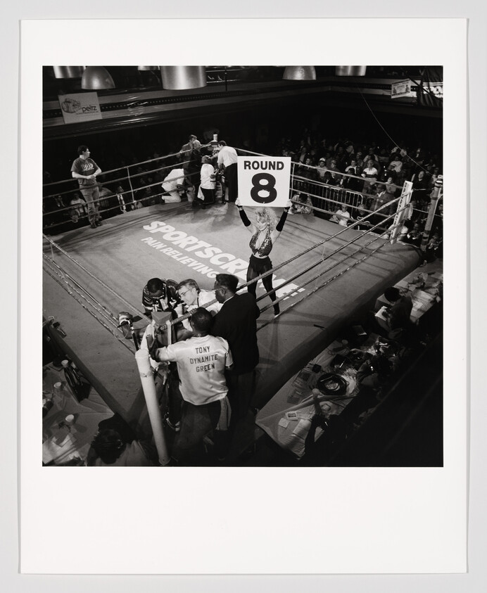 Ring girl holds up a 'Round 8' sign while boxers rest in their corners.