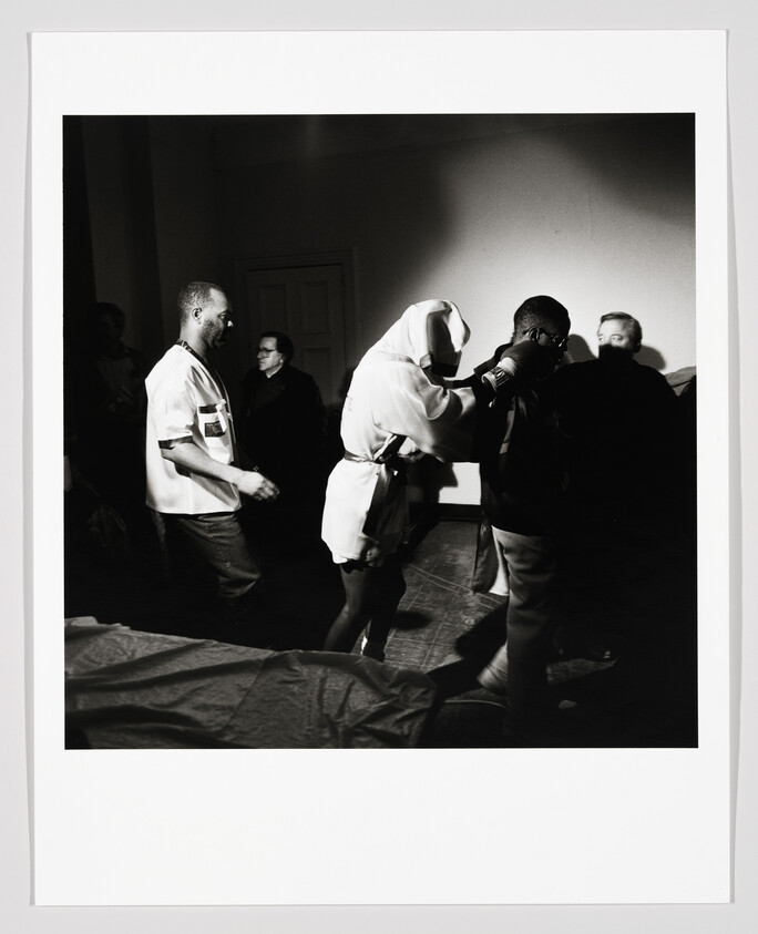 A person in a hooded robe guides a man forward while others watch in a dim room.