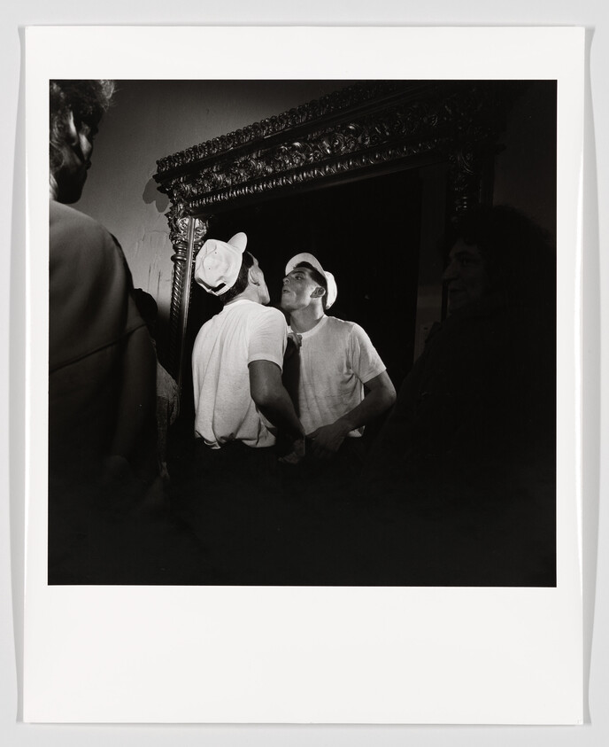 A young man in a white shirt and cap leans forward to kiss his reflection in an ornate mirror.