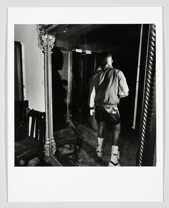 Man in boxing shorts and jacket walks away past a large ornate mirror reflecting his back.