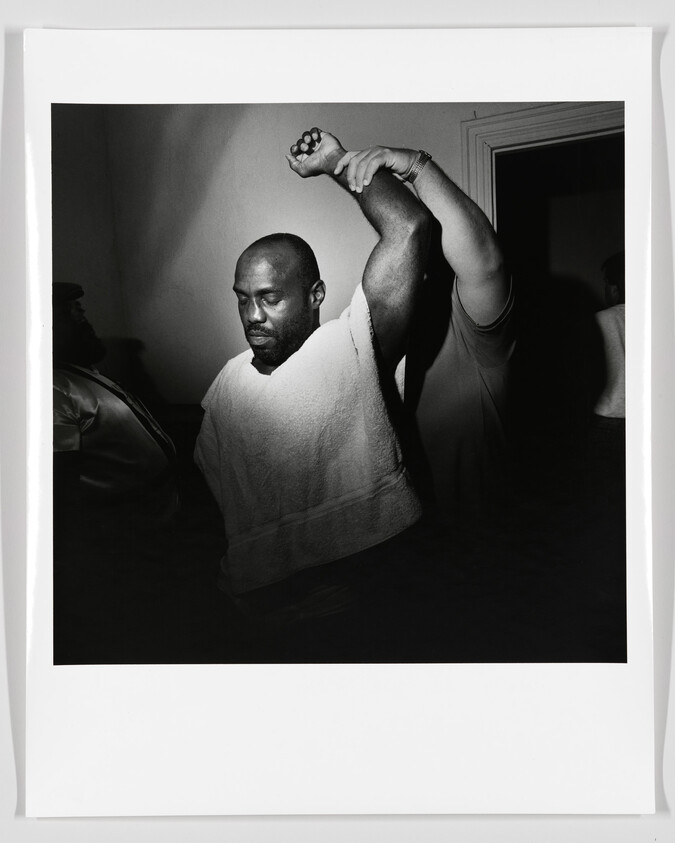 A man with a towel draped over his shoulders holds his arm up while being frisked.