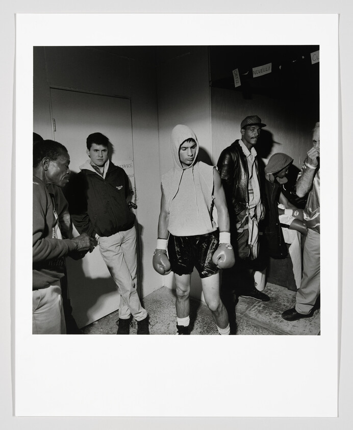 A hooded boxer wearing gloves and shorts walks through a hallway flanked by his team.