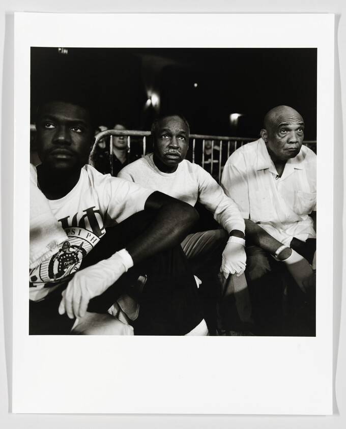 Three men sit side-by-side at a boxing ring, watching the match with serious expressions.
