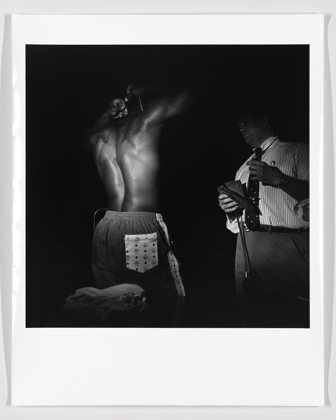 A shirtless athlete has his arms raised while a man in a shirt measures and notes results.