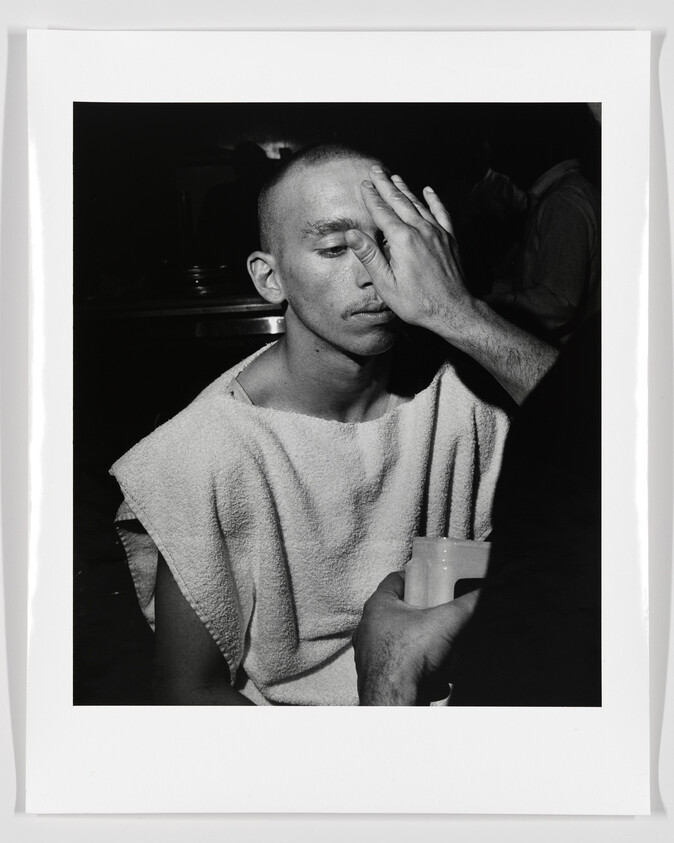 A man with a towel around his shoulders has his face gently pressed by another person's hand.