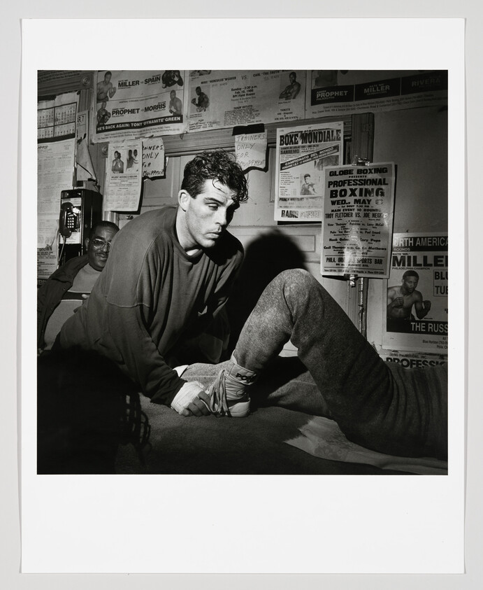 Trainer wraps a boxer's ankle in a dim boxing gym surrounded by vintage fight posters.