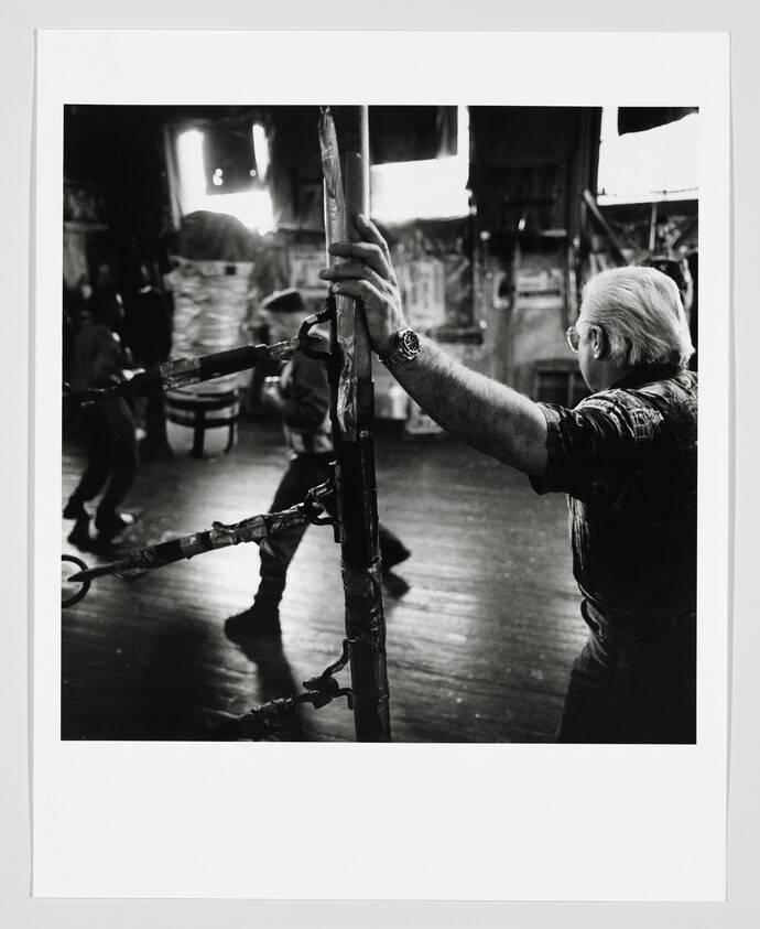 Older man leans on boxing ring ropes watching two fighters spar in a gym.