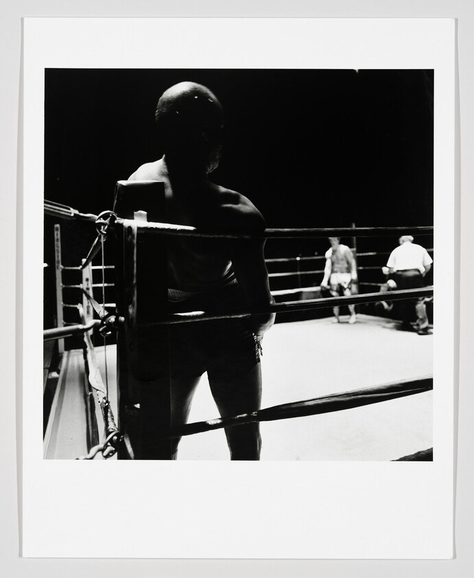 A boxer stands in the corner of the ring watching the opponent and referee prepare.