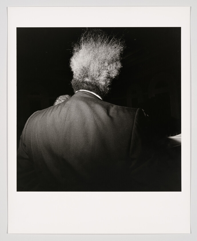 Back of a person in a dark suit with frizzy white hair illuminated against a black background.