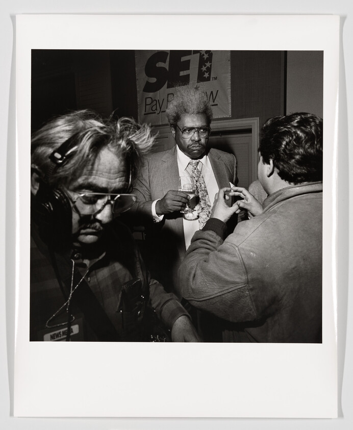 A black and white photograph capturing a busy moment, possibly at an event. In the foreground, a man with headphones and a camera is looking down, seemingly adjusting his equipment. In the center, another man with an afro hairstyle, wearing glasses and a suit, is holding a drink and looking slightly off-camera with a serious expression. To the right, there's a partial view of a person facing the man in the suit, with only the back of their head and shoulder visible. In the background, a sign with the text "SET Pay Per View" is partially visible. The image has a white border, resembling a printed photograph.