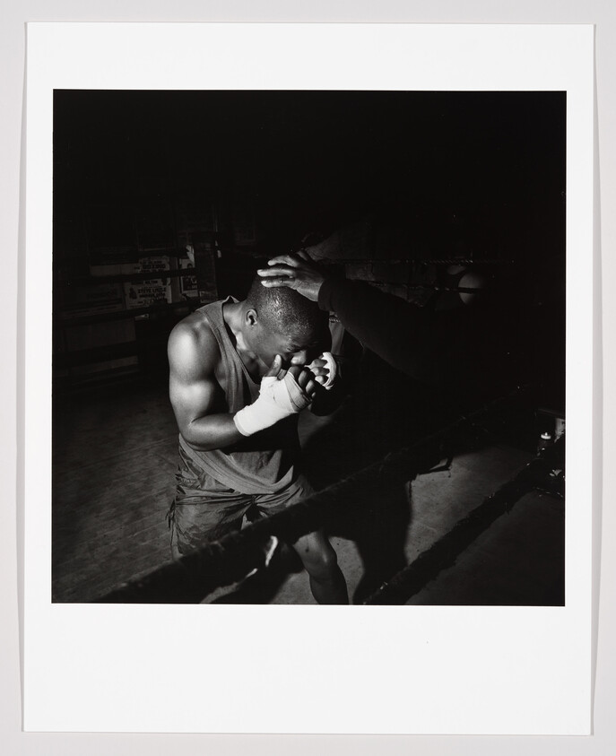 A boxer with wrapped hands crouches in the corner as a trainer steadies his head.