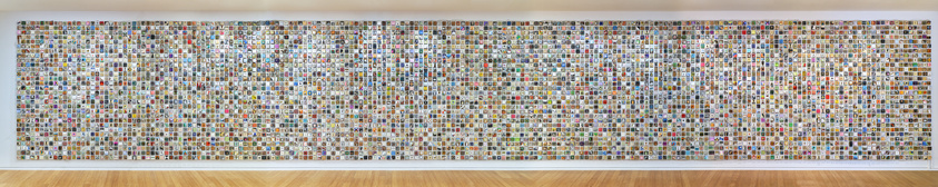 A large wall collage of numerous small, colorful images arranged in a grid pattern, displayed in a gallery with a wooden floor.