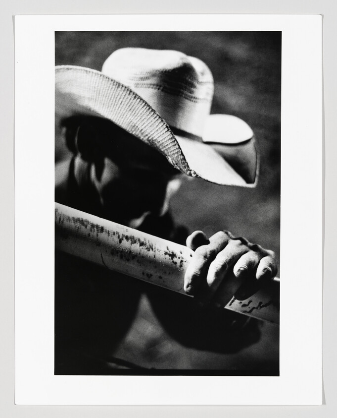A cowboy in a hat grasps a weathered fence rail with his hand.
