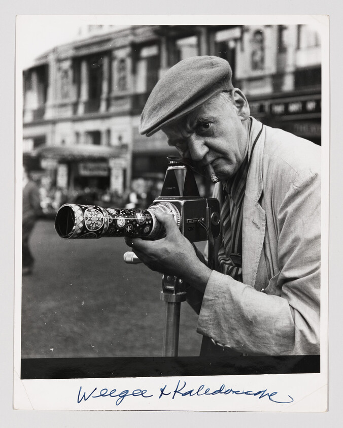 Older man wearing a flat cap looks through a decorated-lens camera mounted on a tripod.