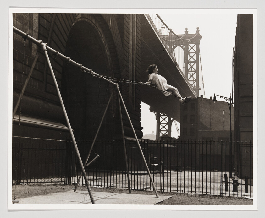 A girl swings high on a playground swing near a large suspension bridge.