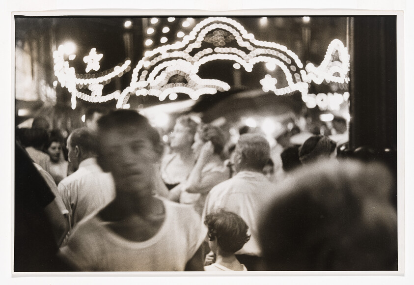 A black and white photograph capturing a bustling street scene at night with blurred figures of people in the foreground and decorative lights forming arches in the background. The lights create a festive atmosphere, and the motion blur suggests the movement of the crowd.