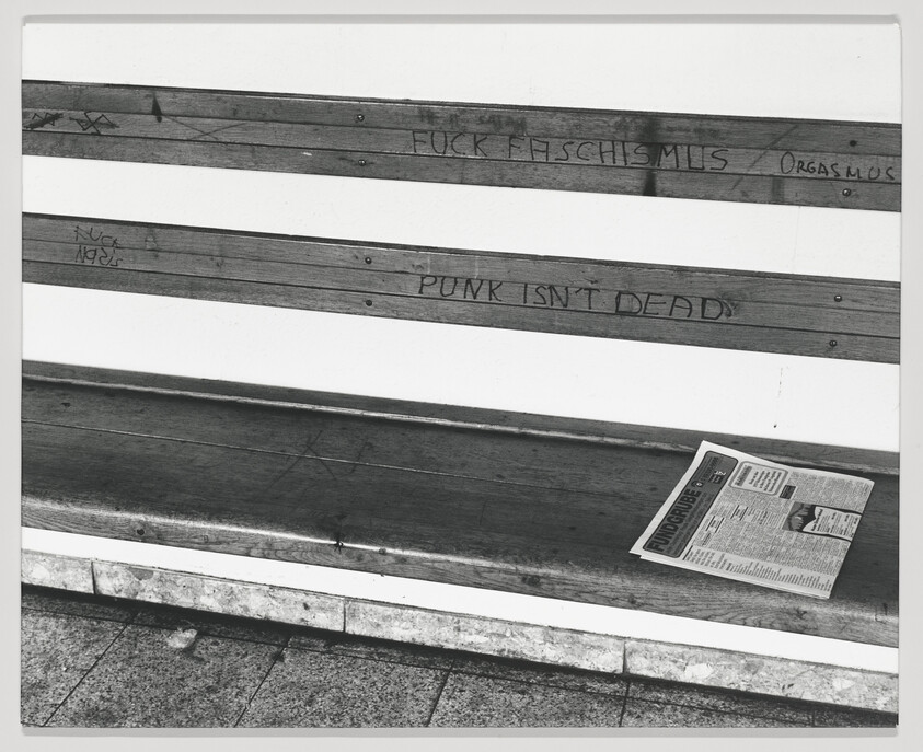 A black and white photograph of a wooden bench with graffiti on it, including the phrase "PUNK ISN'T DEAD," and a newspaper lying on the seat.