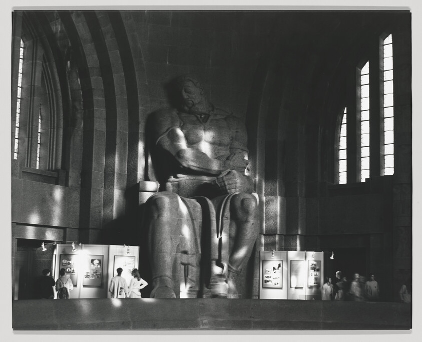A monochrome photograph of a large, seated statue in a hall with high ceilings and tall, narrow windows. The statue appears to be made of stone and depicts a muscular, contemplative figure. Below the statue, there are several display cases with exhibits, and a few visitors are visible, observing the displays or walking through the hall. The lighting casts dramatic shadows and highlights the architectural details of the space.
