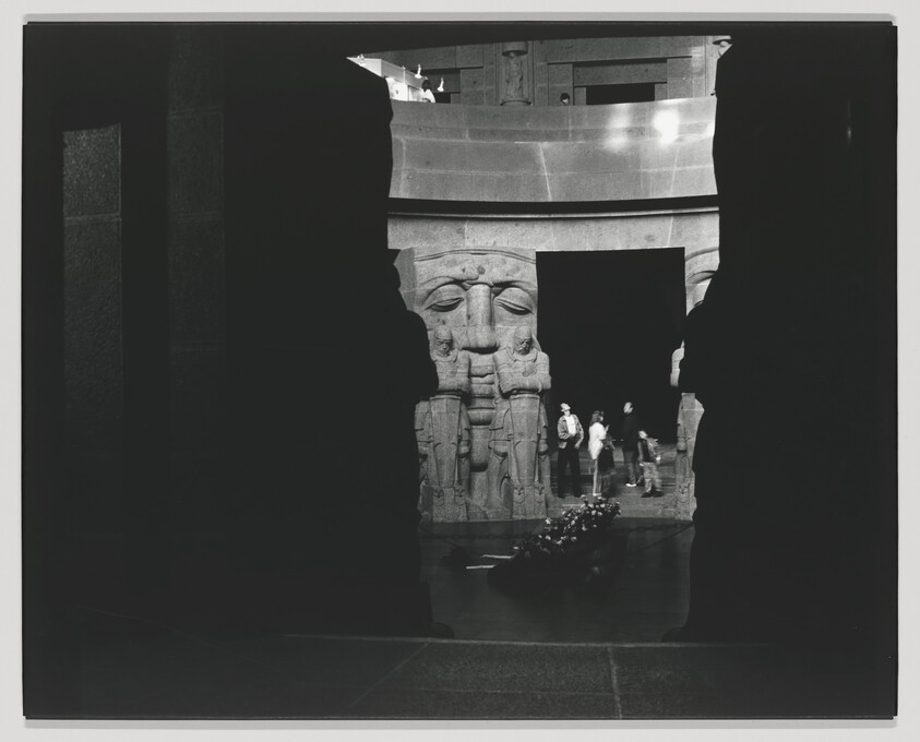 Black and white photo capturing a scene inside what appears to be a museum with an ancient Egyptian artifact, possibly a temple facade with large stone carvings of faces and hieroglyphics. Visitors are seen in the background, some in motion, giving a sense of activity. The artifact is framed by dark silhouettes of architectural elements or statues, creating a dramatic contrast.