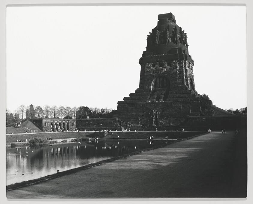 Black and white photograph of the Völkerschlachtdenkmal (Monument to the Battle of the Nations) in Leipzig, Germany, with its imposing structure reflected in a pond in the foreground. People are visible walking around the pond and sitting on the steps of the monument.
