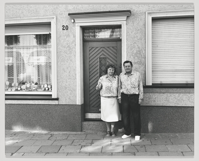 A black and white photo of a middle-aged couple standing in front of a house with the number 20 next to the door. The window to the left displays various trophies, and the window to the right has its shutter closed. The couple is dressed in patterned shirts, and they appear to be posing for the photo.
