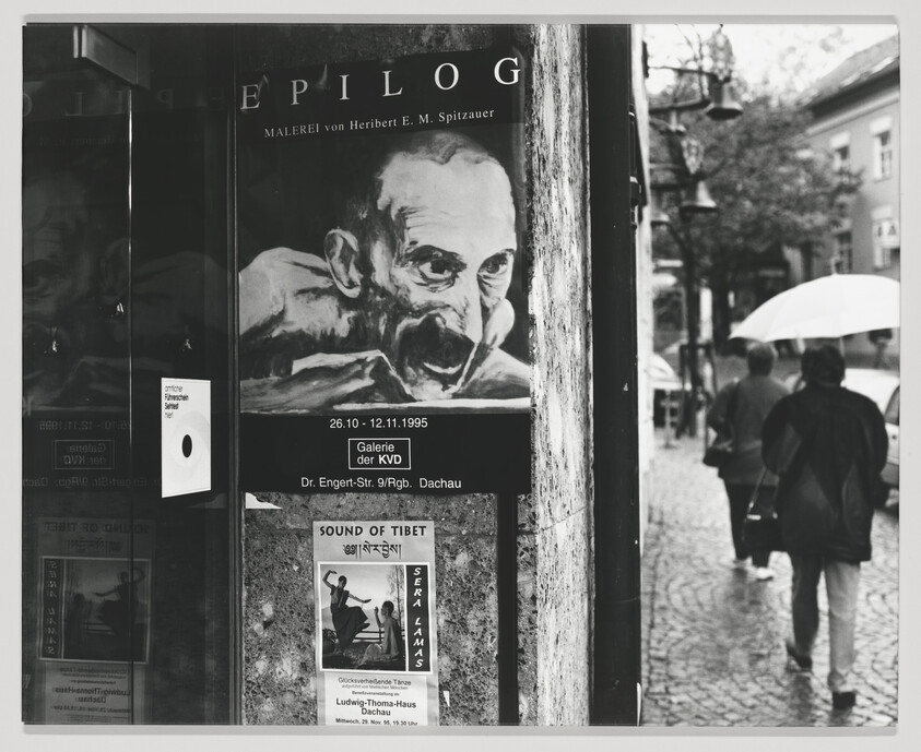 A black and white photograph capturing a street scene with a focus on a public notice board. The board displays posters for cultural events, including an art exhibition titled "EPILOG" featuring paintings by Heribert E. M. Spitzauer, and a performance called "SOUND OF TIBET." In the background, two individuals walk away from the camera, one holding an umbrella, on a cobblestone street lined with street lamps.