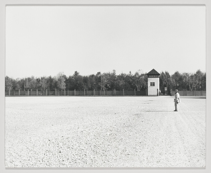 A black and white photograph depicting a vast, empty expanse of gravel with a single person standing in the foreground, looking towards a small guard tower in the distance. The background is lined with a fence and trees under a clear sky.