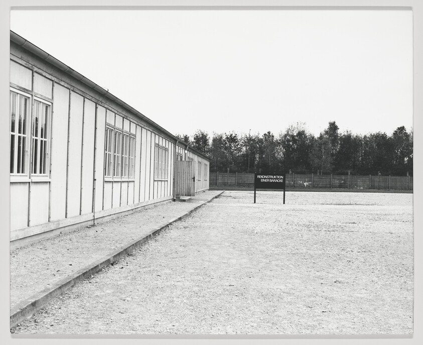 A black and white photograph showing a long barrack building with multiple windows, situated on a barren ground. In the foreground, there's a sign with the text "Rekonstruktion einer Baracke," indicating a reconstruction of a barrack. The background features a fence and a line of trees under a cloudy sky.