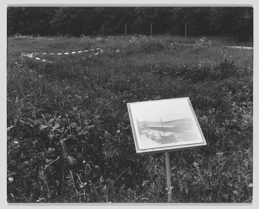 A black and white photograph depicting a grassy field with a historical marker in the foreground. The marker displays an old photograph of an aircraft, possibly from the early days of aviation. In the background, a partial outline of a circle is created by a series of white markers on the ground, suggesting the location of a historical event or object, surrounded by a natural landscape with trees and shrubs.
